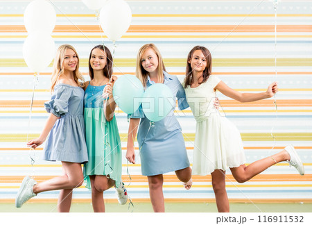 Pretty smiling teenage girls in dresses standing together with ballons at birthday party on the striped colorful background 116911532
