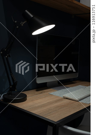 The computer desk is illuminated by a desk lamp in a dark room. The computer desk is illuminated by a desk lamp in a dark room. 116913751