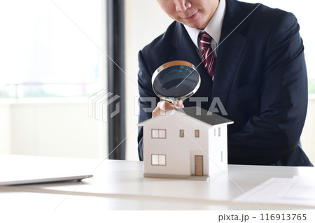 虫眼鏡で住宅模型を覗くスーツ姿の若い男性 116913765