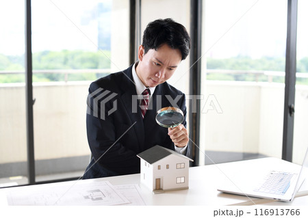 虫眼鏡で住宅模型を覗くスーツ姿の若い男性 116913766
