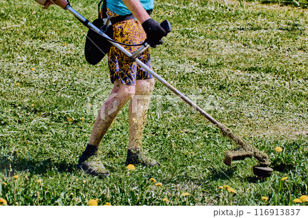 Landscaper legs are covered in shredded grass when he uses weed eater machine without wearing protective clothing. 116913837