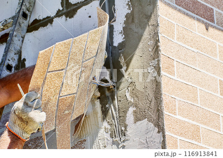 Outdoor facade faux brick wall panel, construction worker makes hole for electrical wiring. Outdoor facade faux brick wall panel, construction worker makes hole for electrical wiring. 116913841