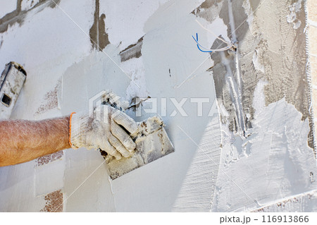 Finishing work on cladding of facade of house, construction worker applies cement based adhesive mortar to walls and levels it with steel spatula. Finishing work on cladding of facade of house, construction worker applies cement based adhesive mortar to walls and levels it with steel spatula. 116913866