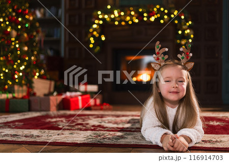 Merry Christmas. Little girl smiling near Christmas tree in classical dark interior. Young happy kid wearing deer horns in living room with fireplace Christmas tree gift boxes. Christmas eve at home Merry Christmas. Little girl smiling near Christmas tree in classical dark interior. Young happy kid wearing deer horns in living room with fireplace Christmas tree gift boxes. Christmas eve at home 116914703