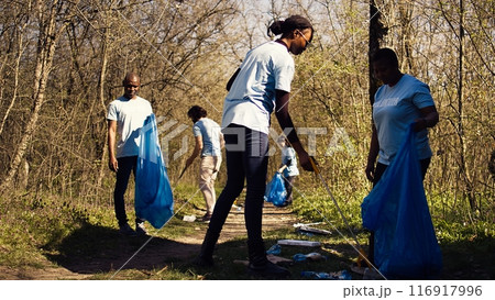 African american team of volunteers picking up trash from the ground, collecting rubbish and recycling in bags. Women cleaning the forest environment from plastic waste, illegal dumping. Camera B. 116917996