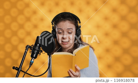 Happy narrator wearing headphones reading aloud from book into mic against yellow background. Joyous lady recording audiobook, creating engaging media content for listeners, camera A 116918078