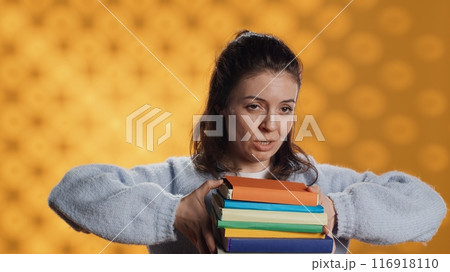 Woman choosing between physical books and ebooks, favoring ereader for lightness, studio background. Bookworm picking digital literature on tablet in front of heavy stack of novels, camera A 116918110