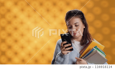 Smiling woman reading messages on smartphone while holding stack of books, isolated over studio background. Cheerful person with pile of academic textbooks in arms texting friends on phone, camera A 116918114