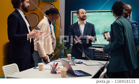 Happy confident coworkers meeting in a conference room to review their results, studying infographics and feedback from stakeholders. Pleased team leader and employees share impressions. Camera B. 116918115