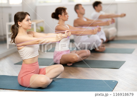 Focused young girl sitting on mat training with resistance band for health wellness, strong biceps or abs muscles during group class in gym health center 116918679