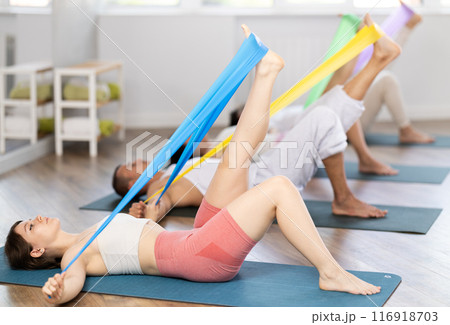 Happy girl and group of women and men lying on mats and doing stretch exercises with resistance bands in fitness room Happy girl and group of women and men lying on mats and doing stretch exercises with resistance bands in fitness room 116918703