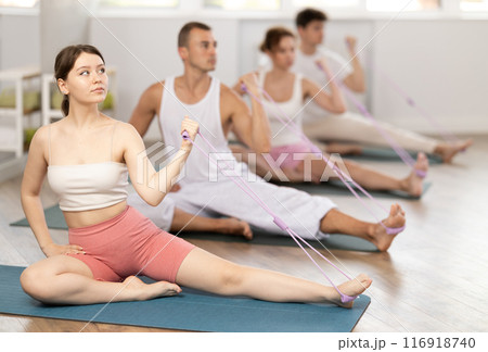 Professional young girl wearing sports clothes using resistance bands during group pilates class sitting on mat in fitness room 116918740