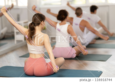 Back view of slim flexible athletic young woman sitting on sports mat practicing pilates during group exercises with elastic band in sports club 116918744