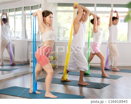 Young men and women doing shoulder workout with resistance band in standing positions Young men and women doing shoulder workout with resistance band in standing positions 116918945