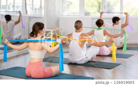 Back view of young girl with group of young adults maintaining physical health practicing pilates with resistance band sitting on gymnastics mat at fitness center 116919005
