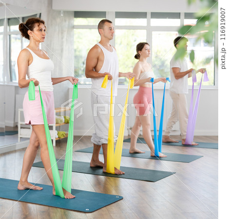 Happy young active woman with group people in sportswear, standing and training exercise with resistance bands in gym 116919007
