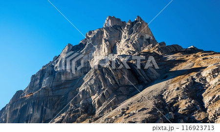 A dramatic, wind-swept peak with sheer cliffs rises against a vibrant blue sky, exuding untamed beauty. 116923713