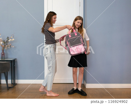 Mother helps put on her daughter's school backpack before school Mother helps put on her daughter's school backpack before school 116924902