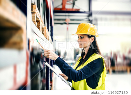 Manager woman in business casual clothes and reflective safety clothes controlling goods in warehouse. 116927180