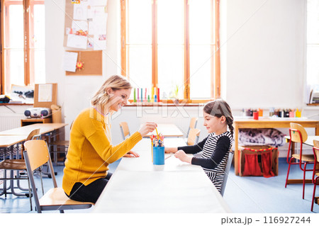Hardworking teacher learning with young schoolgirl in classroom, sitting by desk. Hardworking teacher learning with young schoolgirl in classroom, sitting by desk. 116927244