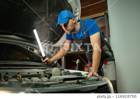 A mechanic wearing a blue shirt and overalls inspects a car engine with a flashlight. He is standing over the engine compartment, looking closely at the engine. A mechanic wearing a blue shirt and overalls inspects a car engine with a flashlight. He is standing over the engine compartment, looking closely at the engine. 116928700