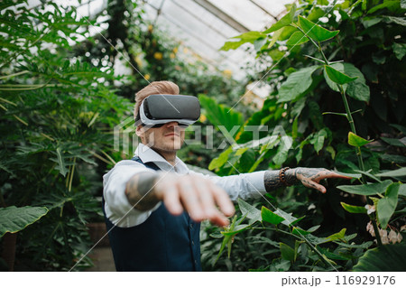 Handsome businessman standing in botanical garden, in the middle of lush green foliage, using VR headset. Handsome businessman standing in botanical garden, in the middle of lush green foliage, using VR headset. 116929176