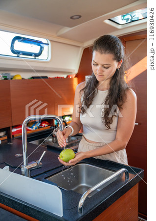 Young woman standing with green apple in kitchen in cabin of modern yacht during sailing in sea. Yacht life concept 116930426