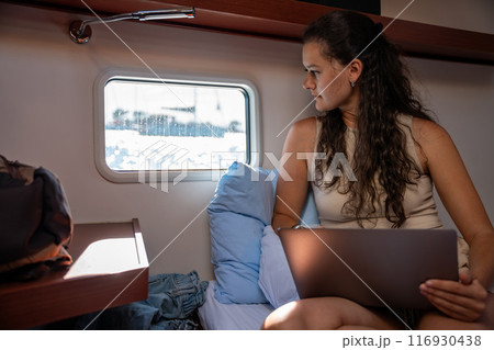 Young woman freelancer using laptop and looking out the window in the cabin of a yacht during sailing in sea. Yacht life concept 116930438
