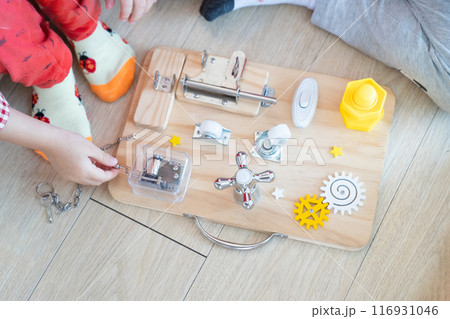 Two Children are playing with busy board toy on the wooden floor together. Children educational busy board toy for developing motor skills. Two Children are playing with busy board toy on the wooden floor together. Children educational busy board toy for developing motor skills. 116931046