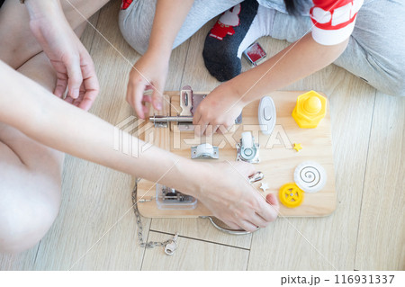 Child and mother are playing with busy board toy on the wooden floor together. Children educational busy board toy for developing motor skills. 116931337