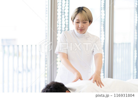 ケーシー白衣を着て施術を行う金髪ショートヘアの若い女性整体師（髪色自由、自分らしく働く、職場環境） 116931804