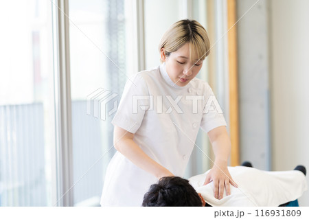 ケーシー白衣を着て施術を行う金髪ショートヘアの若い女性整体師（髪色自由、自分らしく働く、職場環境） 116931809