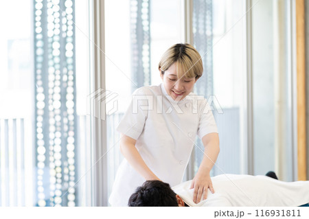 ケーシー白衣を着て施術を行う金髪ショートヘアの若い女性整体師（髪色自由、自分らしく働く、職場環境） 116931811