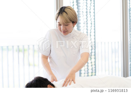 ケーシー白衣を着て施術を行う金髪ショートヘアの若い女性整体師(髪色自由、自分らしく働く、職場環境) ケーシー白衣を着て施術を行う金髪ショートヘアの若い女性整体師(髪色自由、自分らしく働く、職場環境) 116931813
