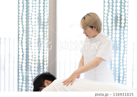 ケーシー白衣を着て施術を行う金髪ショートヘアの若い女性整体師（髪色自由、自分らしく働く、職場環境） 116931815