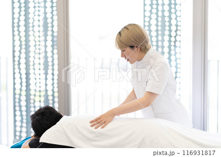 ケーシー白衣を着て施術を行う金髪ショートヘアの若い女性整体師（髪色自由、自分らしく働く、職場環境） 116931817