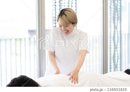 ケーシー白衣を着て施術を行う金髪ショートヘアの若い女性整体師（髪色自由、自分らしく働く、職場環境） 116931820