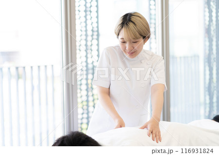 ケーシー白衣を着て施術を行う金髪ショートヘアの若い女性整体師（髪色自由、自分らしく働く、職場環境） 116931824