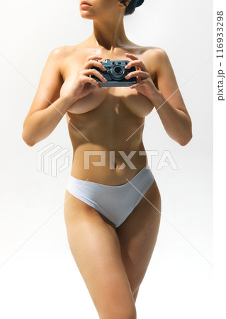 Cropped image of young women with slim tanned body, posing naked with vintage camera against light background. 116933298
