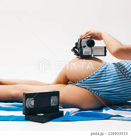 Back view, cropped image of woman wearing striped swimsuit, lying on beach towel with vintage camera and cassette. Memories Back view, cropped image of woman wearing striped swimsuit, lying on beach towel with vintage camera and cassette. Memories 116933313