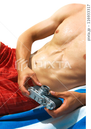 Summer memories. Cropped image of tanned, muscular male body, torso, model in red swim shorts lying on beach towel with vintage camera Summer memories. Cropped image of tanned, muscular male body, torso, model in red swim shorts lying on beach towel with vintage camera 116933317