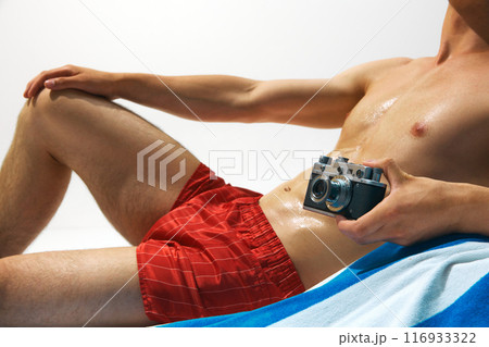 Cropped image of tanned, muscular male body, torso, model in red swim shorts lying on beach towel with vintage camera Cropped image of tanned, muscular male body, torso, model in red swim shorts lying on beach towel with vintage camera 116933322