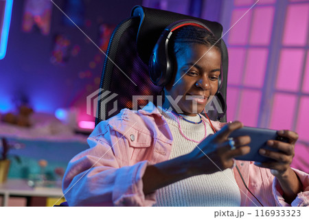 Portrait of teenage African American girl playing mobile games while relaxing at home at night Portrait of teenage African American girl playing mobile games while relaxing at home at night 116933323