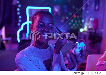 Portrait of smiling young Black woman testing luminescent makeup in dark room with neon lights, copy space 116933349