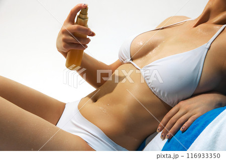 Cropped image of tanned young woman using spf spray for healthy sunbathing, model wearing white swimsuit and lying on beach towel. 116933350