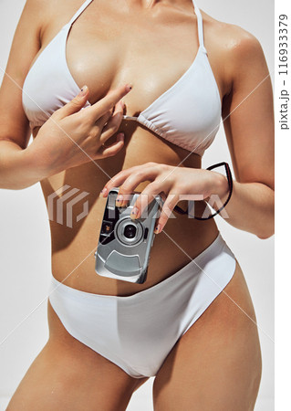 Sensual memories. Cropped image of female body, woman wearing white swimsuit, posing with retro camera against light background 116933379
