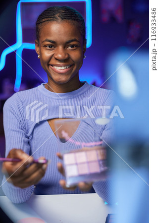 Vertical portrait of smiling young woman recording beauty video and doing make up tutorial holding eyeshadow palette 116933486