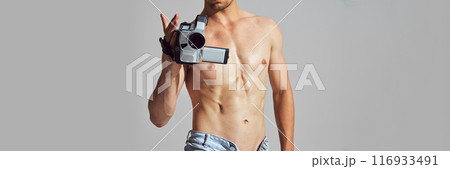 Retro Tech. Cropped image of shirtless man with muscular, athletic body, in denim short standing with vintage camcorder and filming 116933491