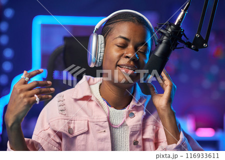 Portrait of inspired African American girl singing to microphone and recording music in home studio with pink neon lights copy space 116933611