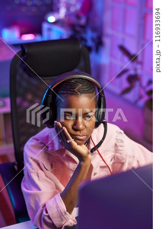 Vertical portrait of young African American woman wearing headphones and watching videos or podcast online with bored face expression 116933694